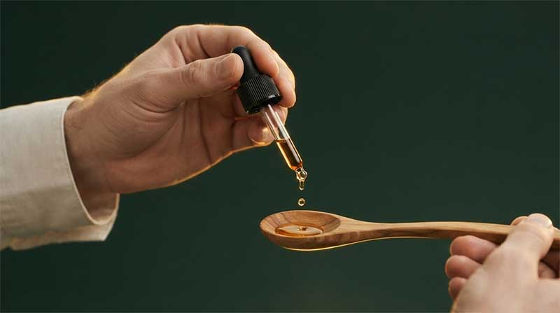 A hand pouring dark black seed oil from a small glass bottle onto a wooden spoon