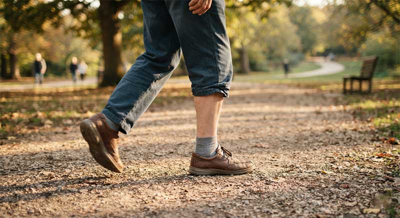An older man's legs and feet walking on a gravel path in a park