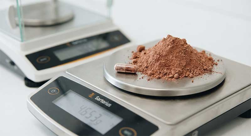 A small supplement capsule next to a much larger pile of loose powder on a lab scale