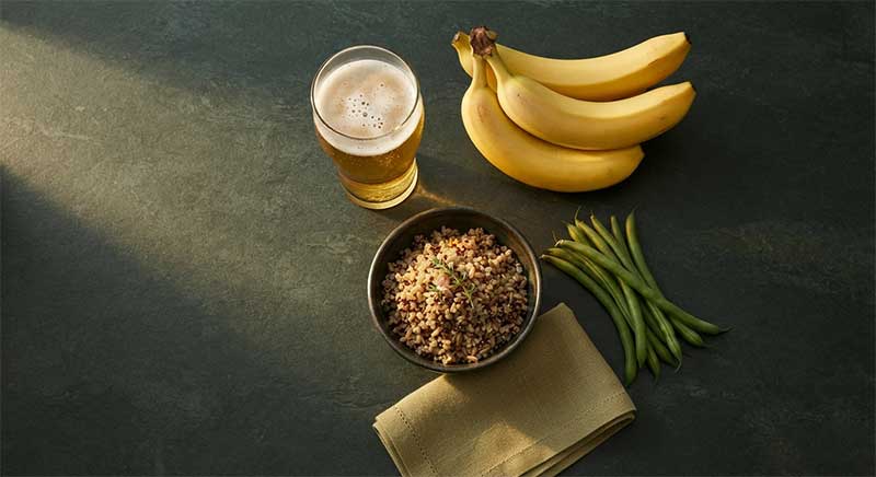 A glass of beer, bananas, green beans, and whole grains on a dark slate surface