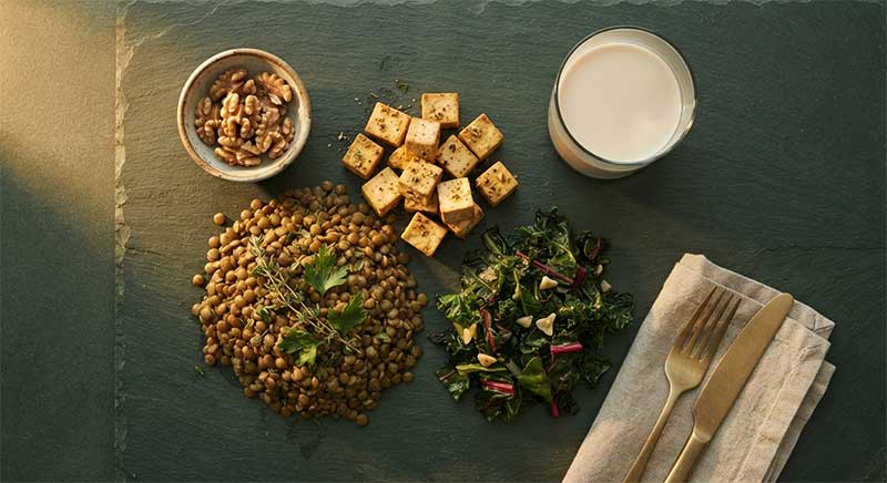 Cooked lentils, tofu, fortified plant milk, leafy greens, and walnuts on a dark slate surface