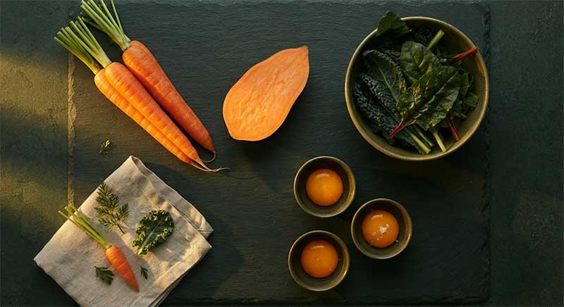 Carrots, sweet potato, dark leafy greens, and egg yolks on a dark slate surface